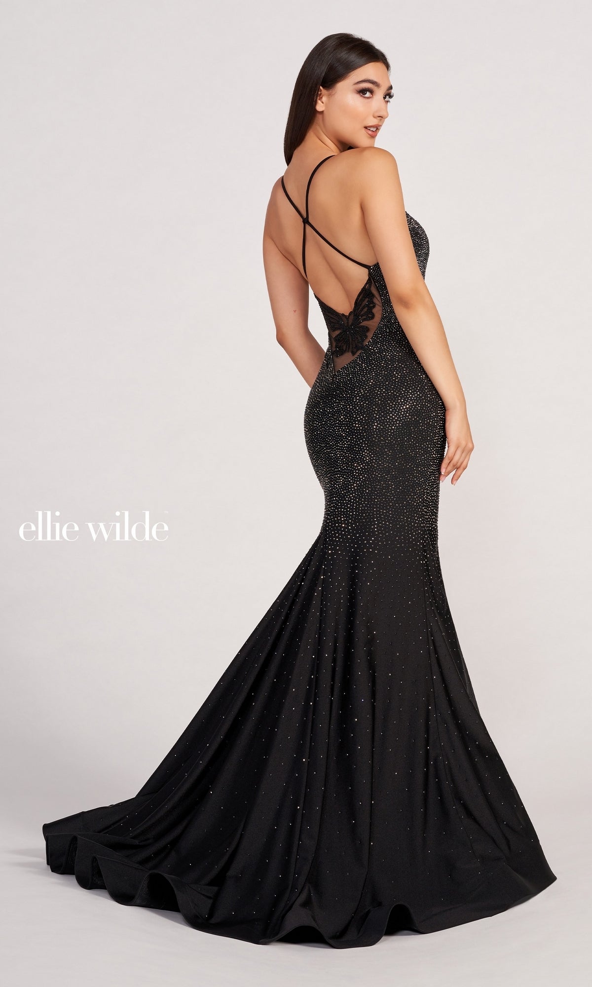 A woman in an Ellie Wilde Butterfly-Back Long Beaded Prom Dress featuring sparkling embellishments and crisscross butterfly straps stands against a plain background.